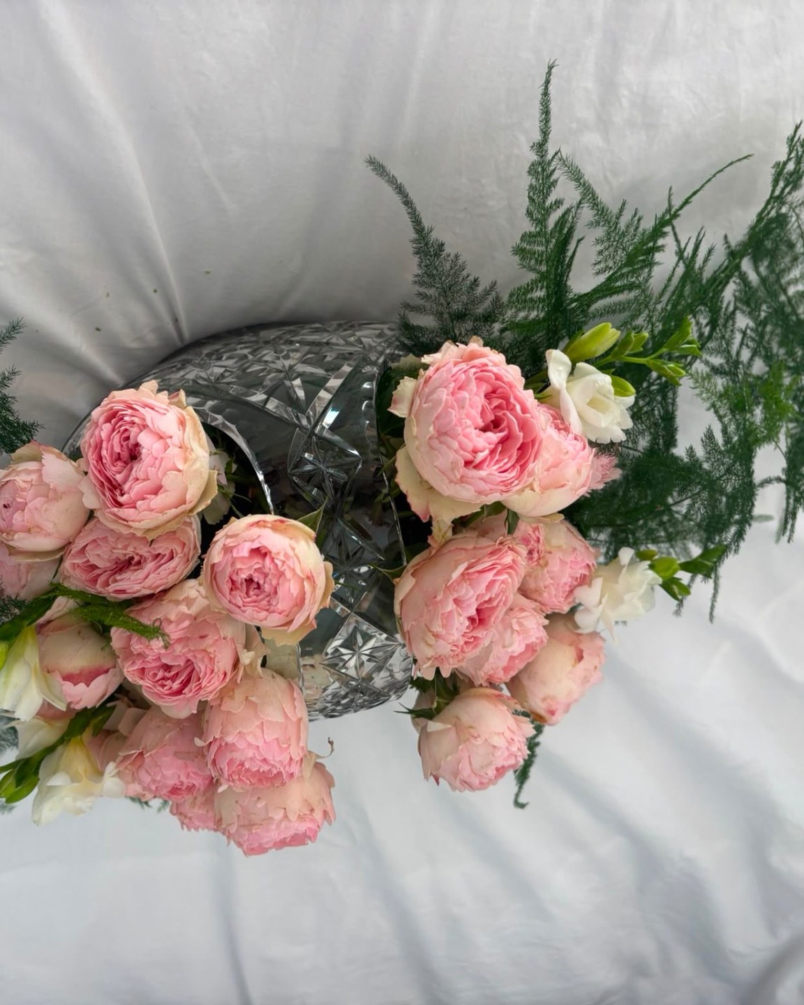 Centrepiece of pink garden roses, peonies, and white freesias with greenery in a cut-crystal vase